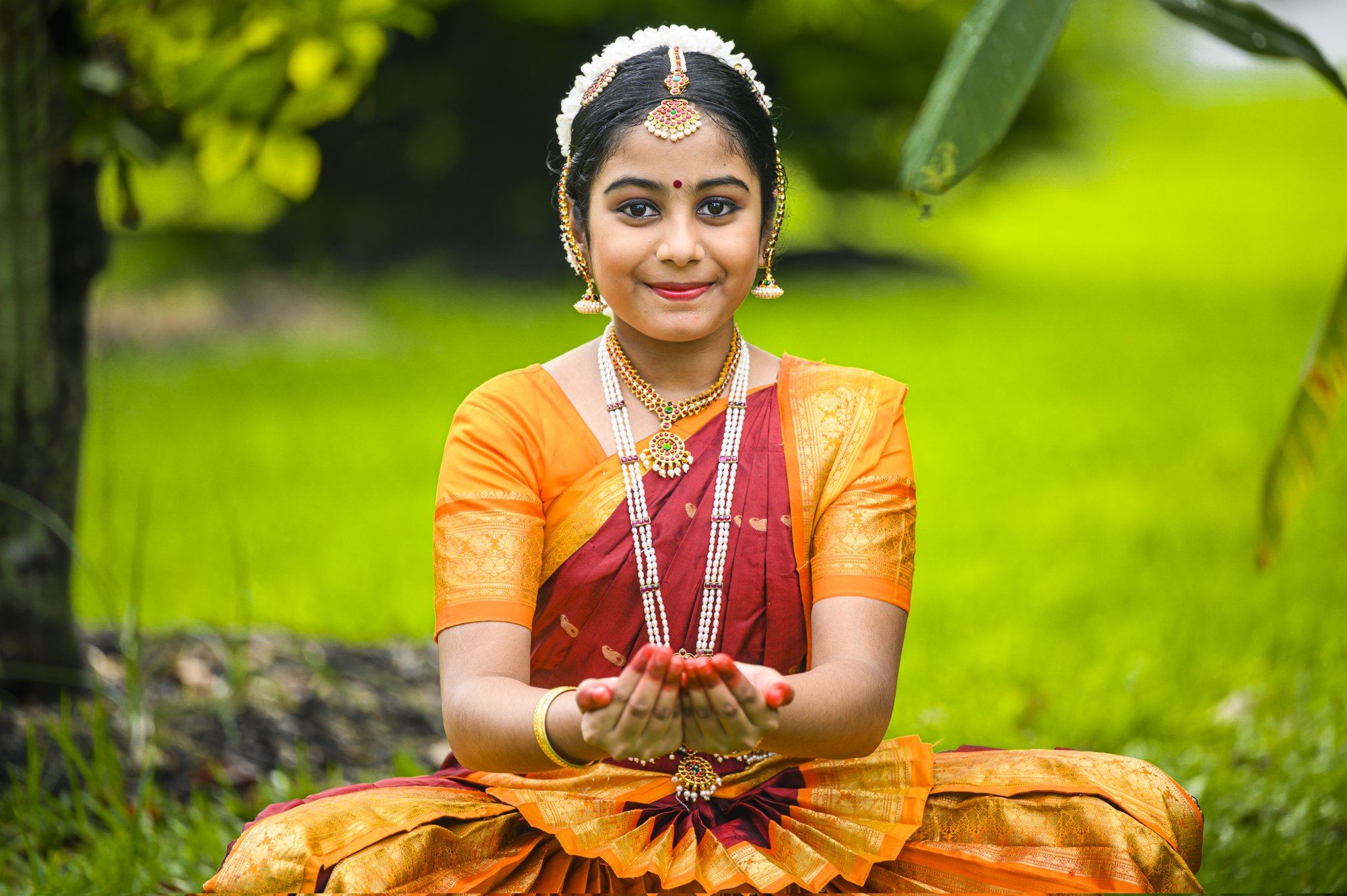 A young dancer seated in Bharatanatyam pose, smiling, with hands cupped